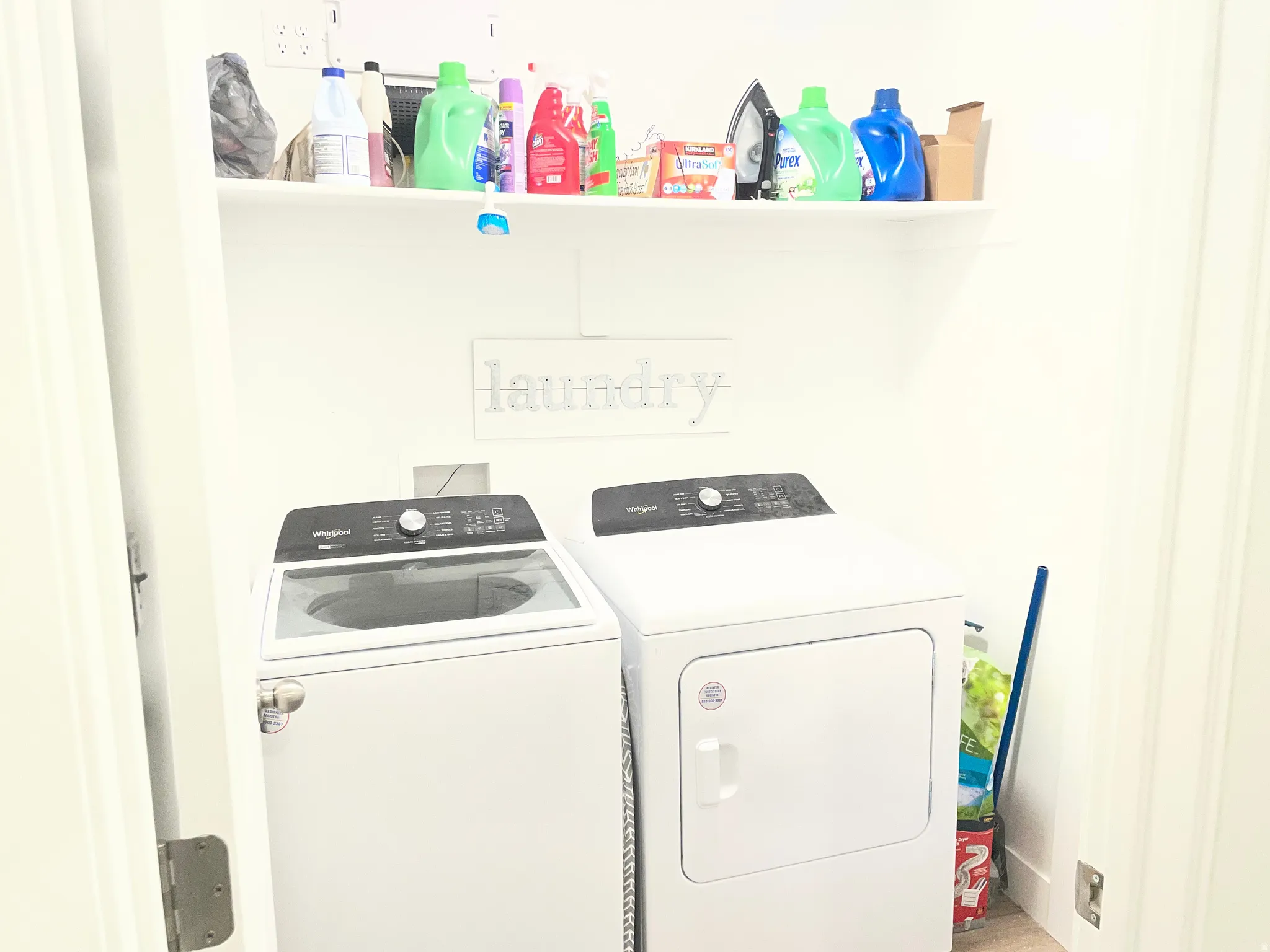 Dedicated laundry room featuring side-by-side washer and dryer with digital controls, white cabinetry, and open shelving for storage of cleaning supplies and household essentials