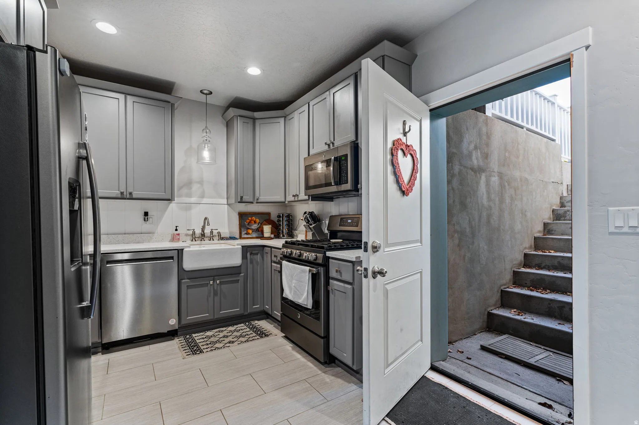 Stainless steel appliances, gray shaker-style cabinetry, white farmhouse sink, light tile flooring, and recessed lighting