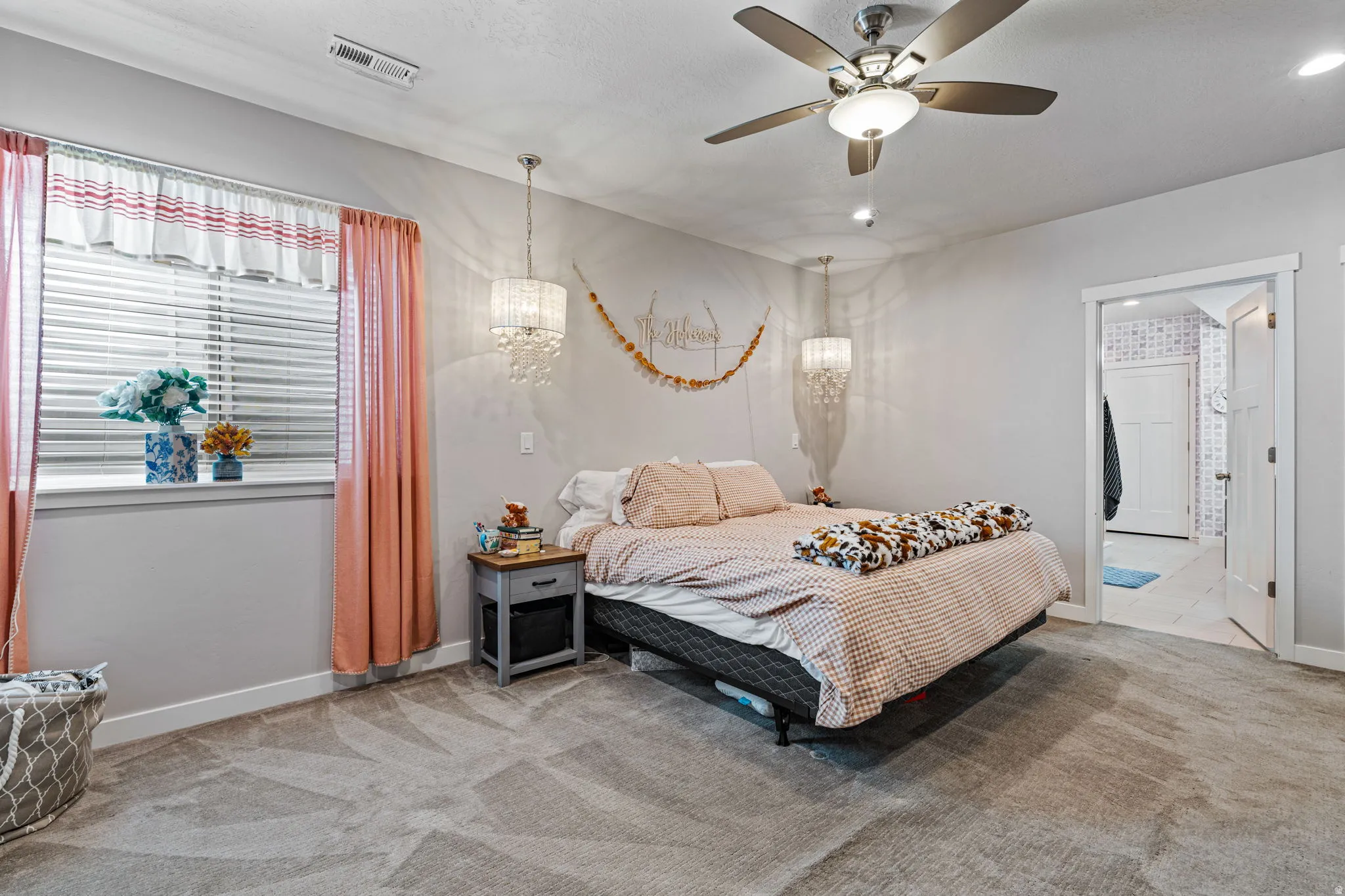 Spacious primary bedroom featuring neutral décor, carpeted flooring, a ceiling fan with integrated lighting, and abundant natural light from a window with coral curtains