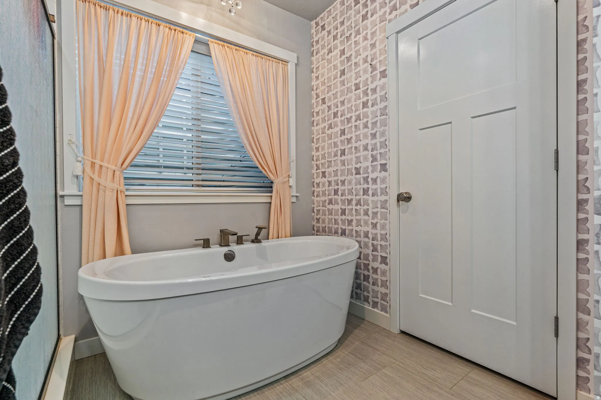 Freestanding soaking tub with dark-toned fixtures, window with horizontal blinds, patterned wall covering, three-panel interior door, and light-toned wood-finish flooring
