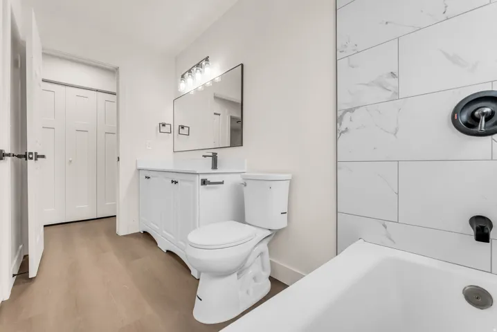 Bathroom featuring a white vanity with a dark faucet, rectangular mirror with overhead lighting, wood-finish flooring, and white tiled shower surround