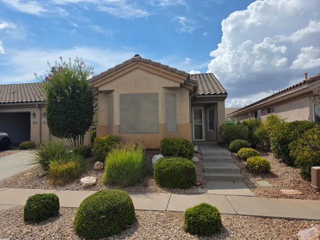 Stucco exterior with a tiled roof, featuring a concrete walkway, mature landscaping, and an integrated garage