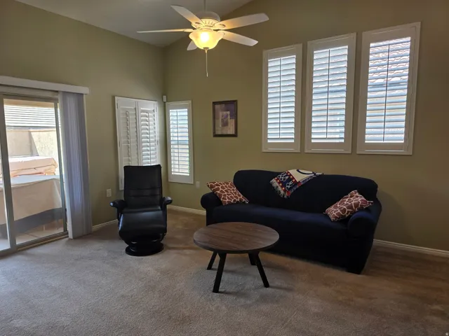 Sliding glass door leading to an outdoor space, high vaulted ceiling with a ceiling fan, multiple windows with white plantation shutters, and light-colored carpet flooring