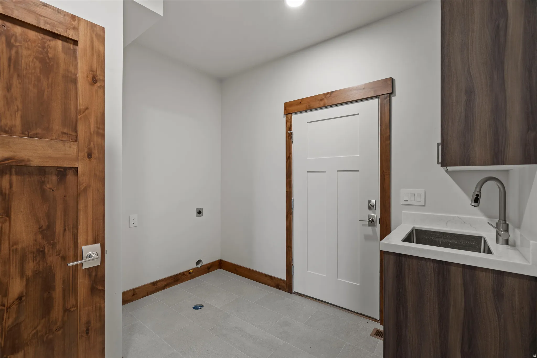 Modern utility space featuring concrete flooring, custom wood-trimmed architectural details, stainless steel sink with contemporary faucet, dark wood-finish cabinetry, white six-panel entry door, and recessed lighting
