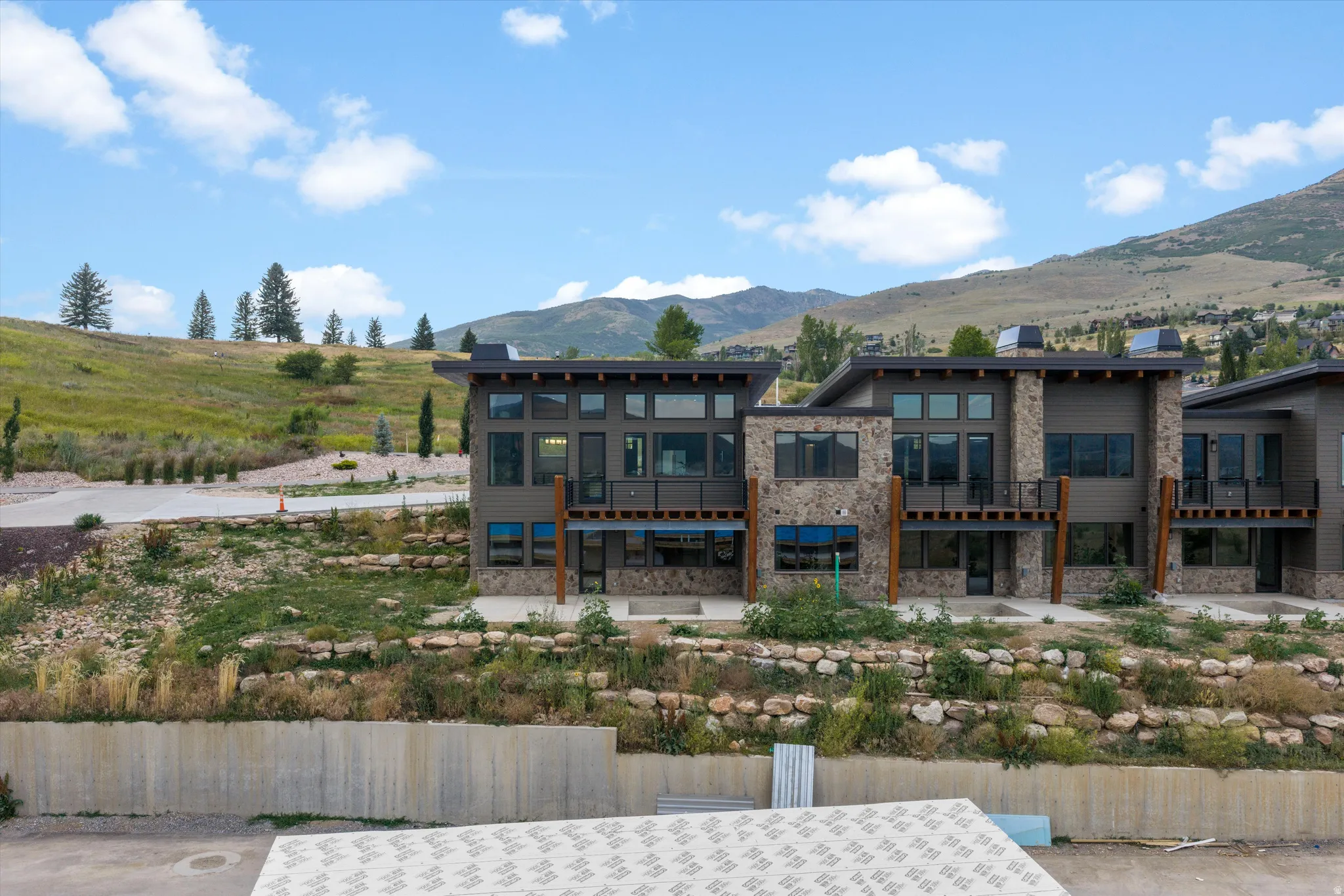 Modern multi-unit residential complex featuring contemporary architecture with mixed material facades of stone and metal cladding, expansive windows with blue-tinted glazing, and tiered landscaping with native plantings and retaining walls
