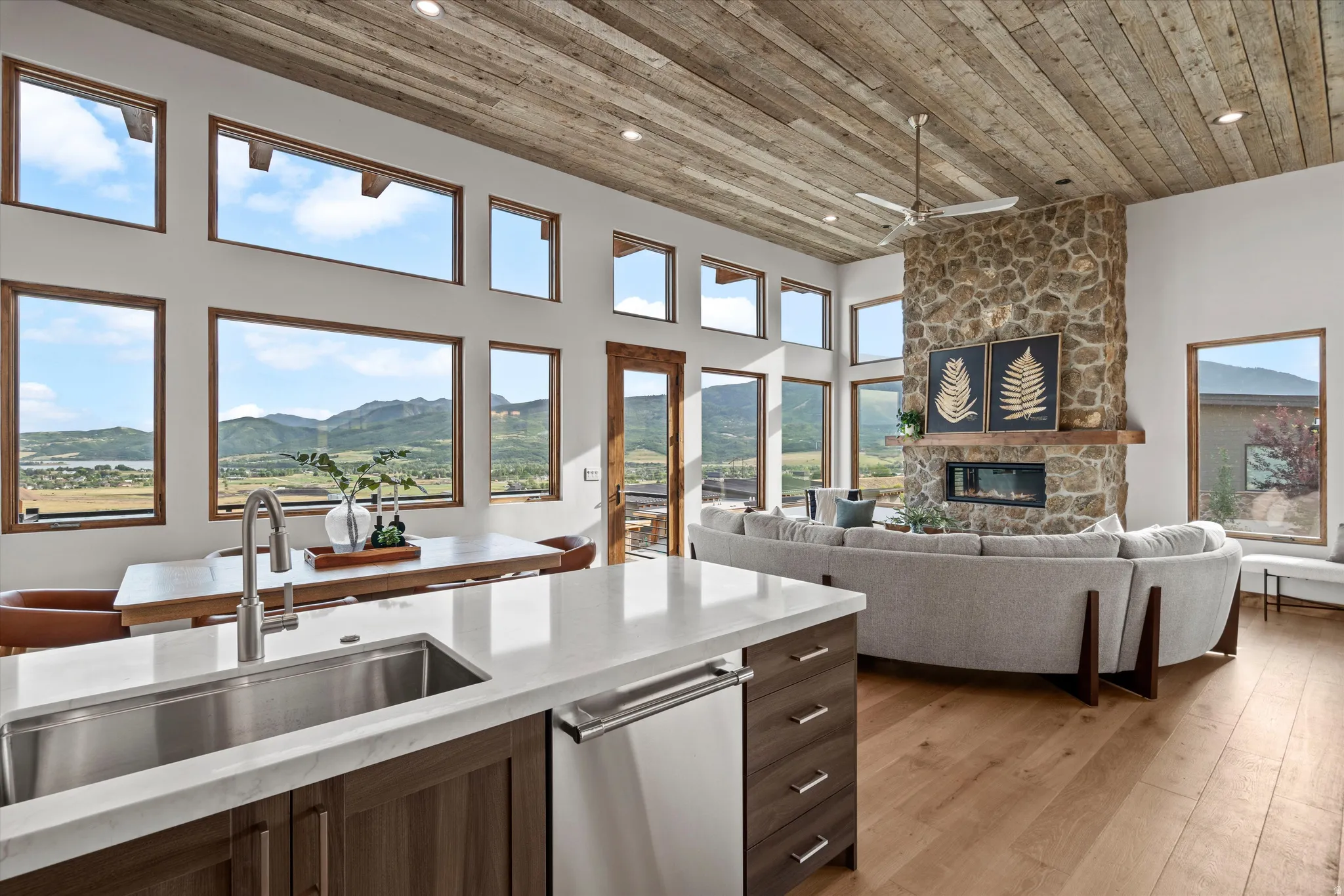 Open-concept living space showcasing floor-to-ceiling windows framing expansive mountain and valley vistas, a wood-finish plank ceiling, stainless steel kitchen island with waterfall countertop, stone fireplace accent wall, and hardwood flooring throughou