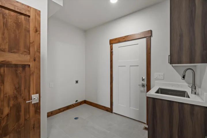 Modern utility space featuring concrete flooring, custom wood-trimmed architectural details, stainless steel sink with contemporary faucet, dark wood-finish cabinetry, white six-panel entry door, and recessed lighting
