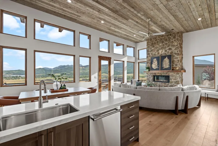 Open-concept living space showcasing floor-to-ceiling windows framing expansive mountain and valley vistas, a wood-finish plank ceiling, stainless steel kitchen island with waterfall countertop, stone fireplace accent wall, and hardwood flooring throughou