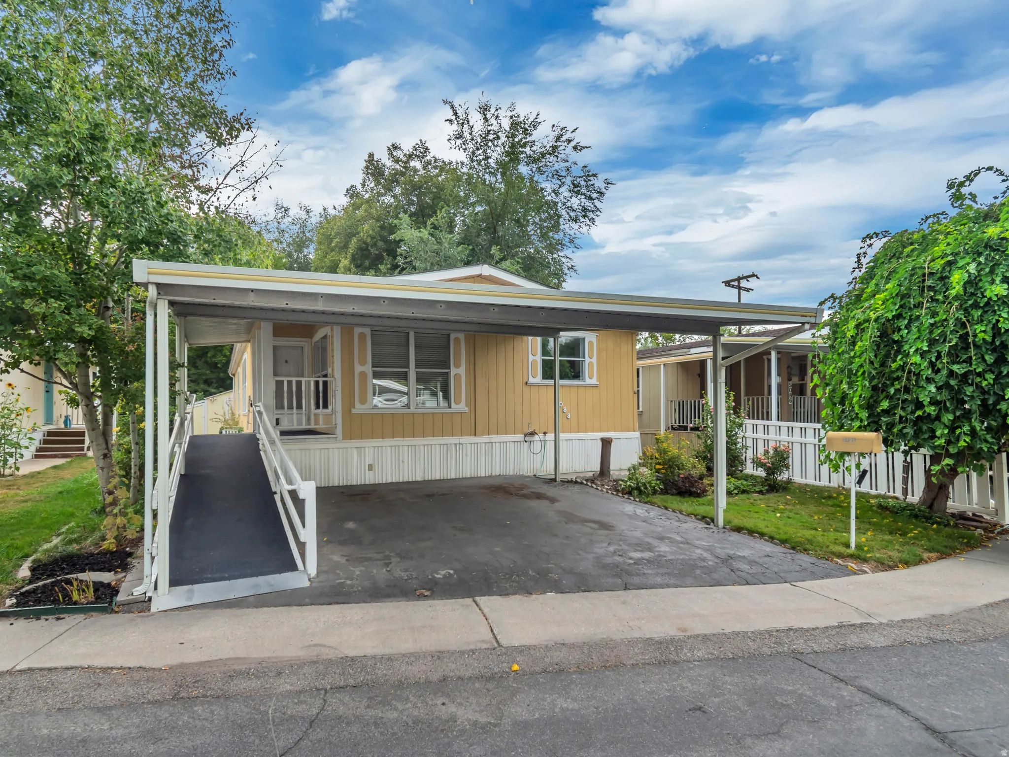 Carport with asphalt driveway, ramp access to entry, exterior siding, and established landscaping Surrounded by trees.