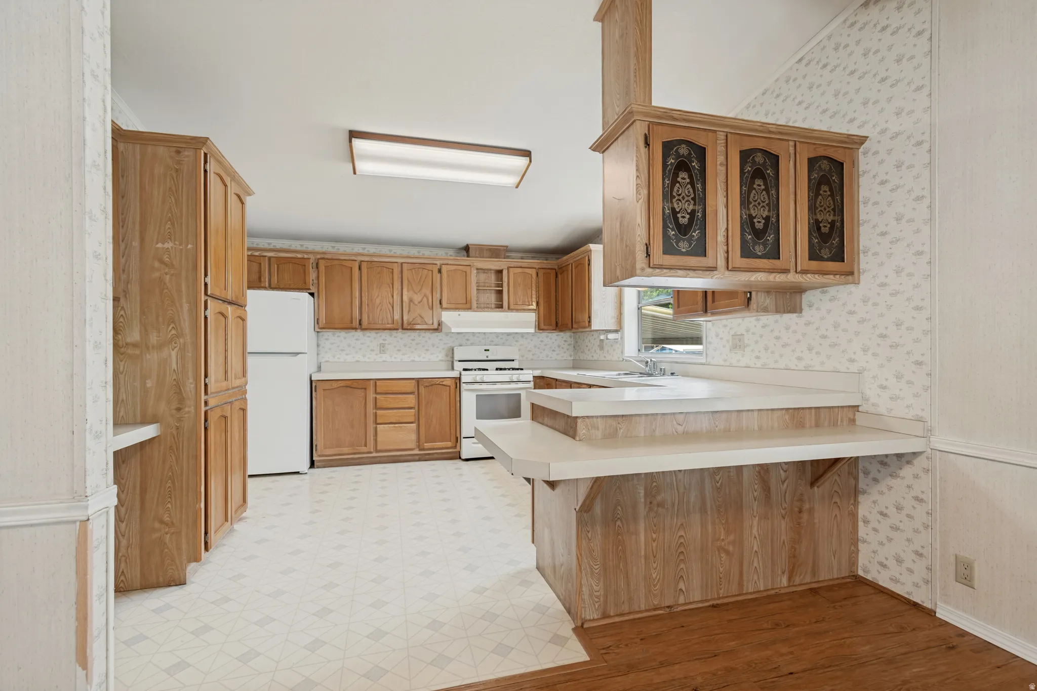 Bright open kitchen featuring wood-finish cabinetry, white appliances, and a built-in breakfast bar