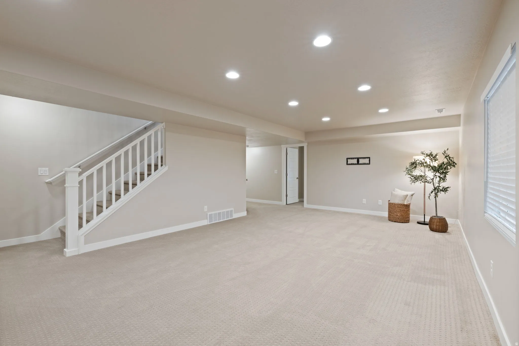Spacious room with recessed lighting, light-toned carpeting, a white balustrade staircase, and a window with blinds