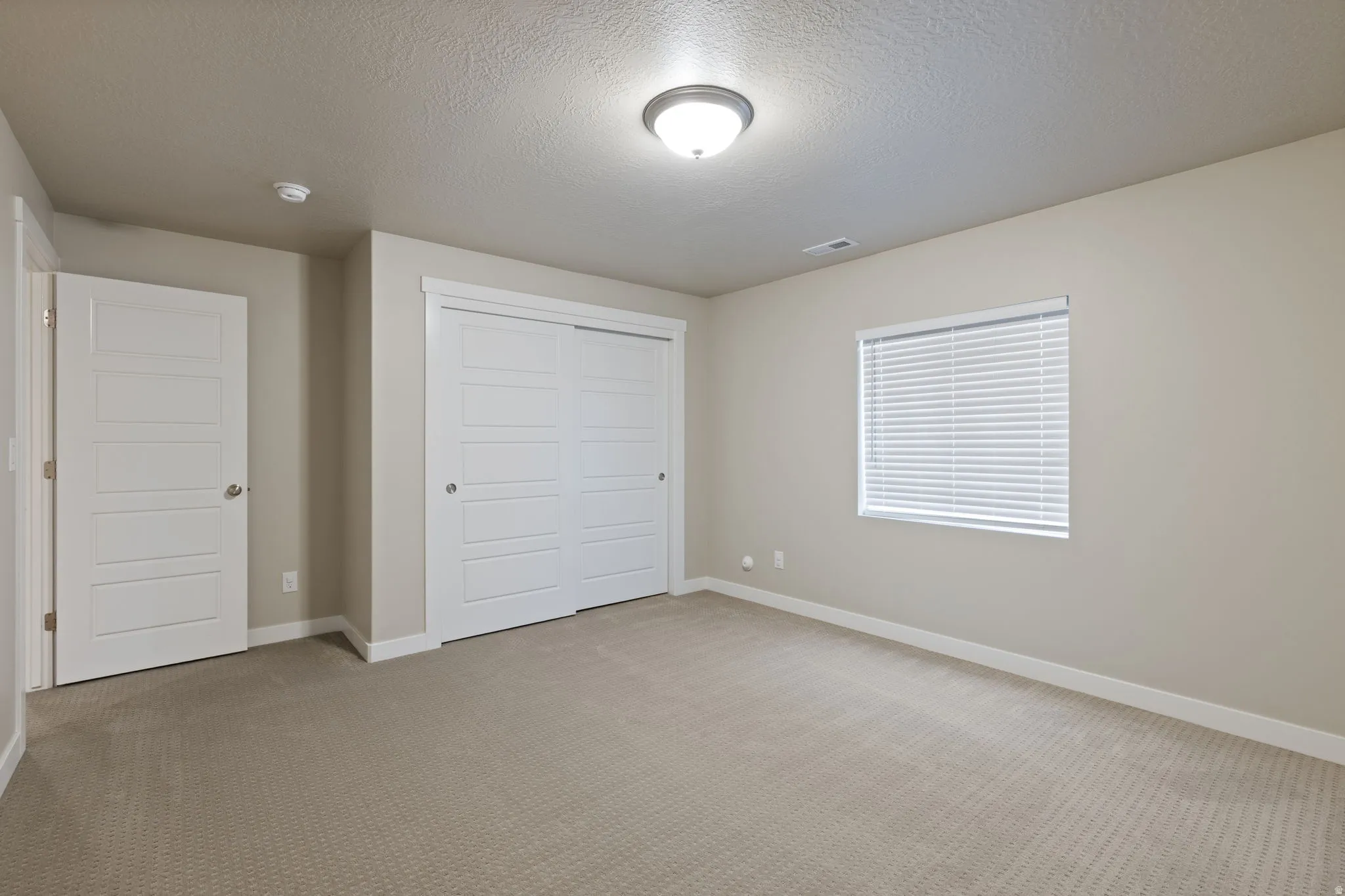 Neutral tone wall paint, carpet flooring, a window with blinds, a sliding closet door, and a flush mount ceiling light