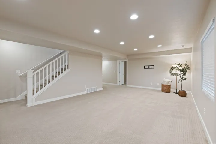 Spacious room with recessed lighting, light-toned carpeting, a white balustrade staircase, and a window with blinds