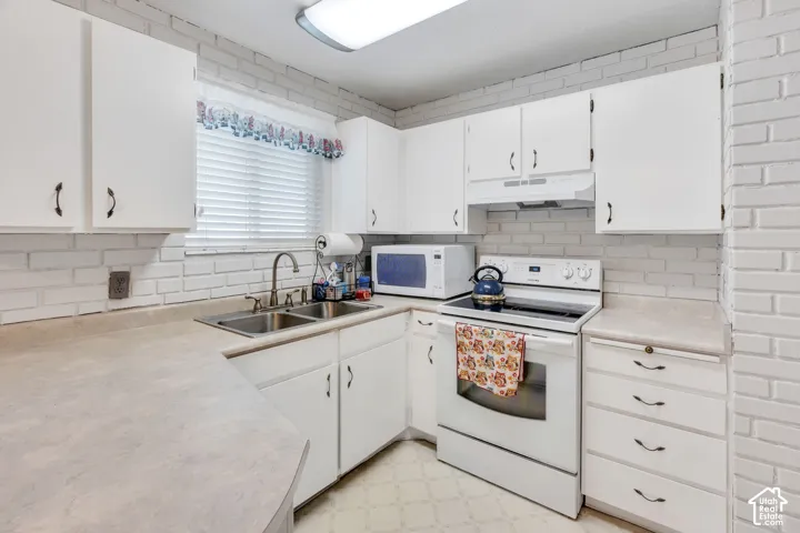 White cabinetry with dark hardware, dual basin stainless steel sink, light-toned countertops, painted brick backsplash, and white range
