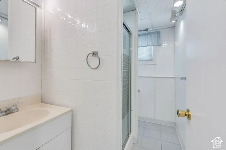 White tiled walls, single vanity with integrated sink, chrome faucet, wall-mounted mirror, and glass-enclosed shower