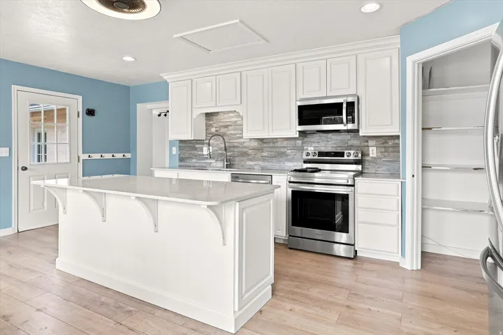 Kitchen featuring white cabinetry, stainless steel appliances, a stone tile backsplash, a center island with a light-toned countertop, and wood-finish flooring