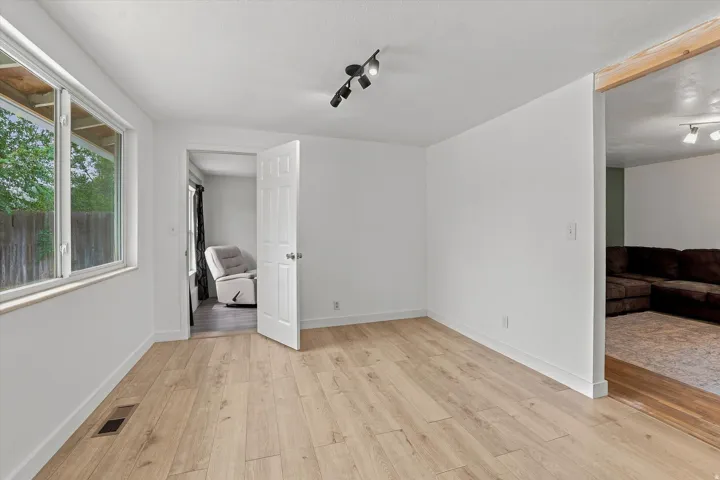 Light wood-finish flooring, white walls, large window with white frame, track lighting, and a white paneled interior door
