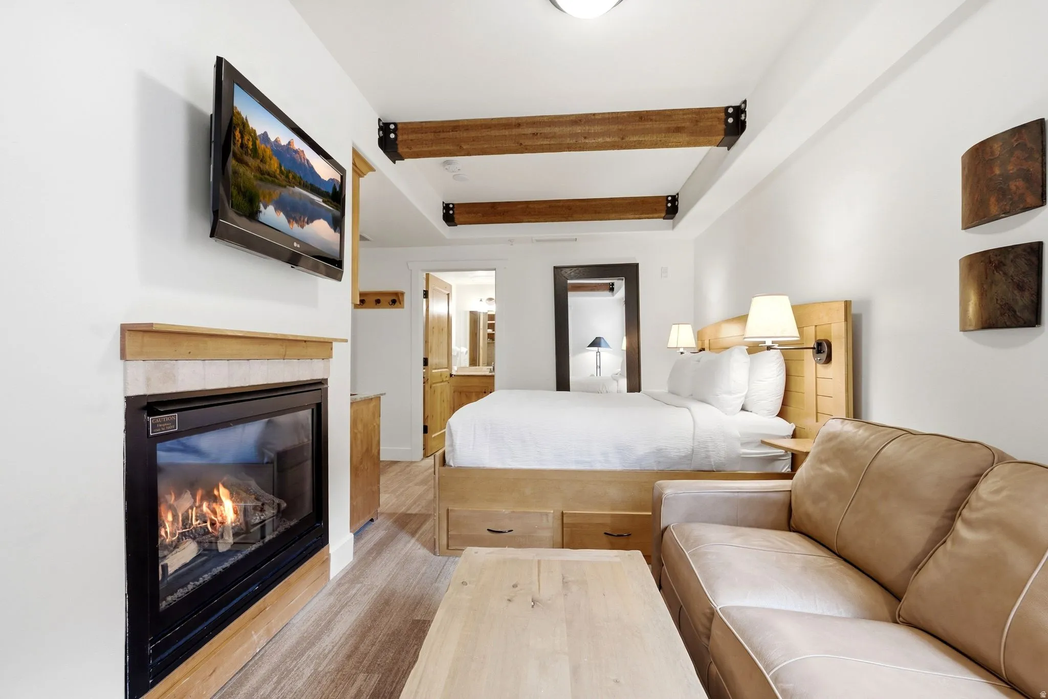 Wood-finish flooring, built-in fireplace with tile surround, exposed wood beams, wood panel bed frame with integrated nightstands, and wall-mounted sconce lighting