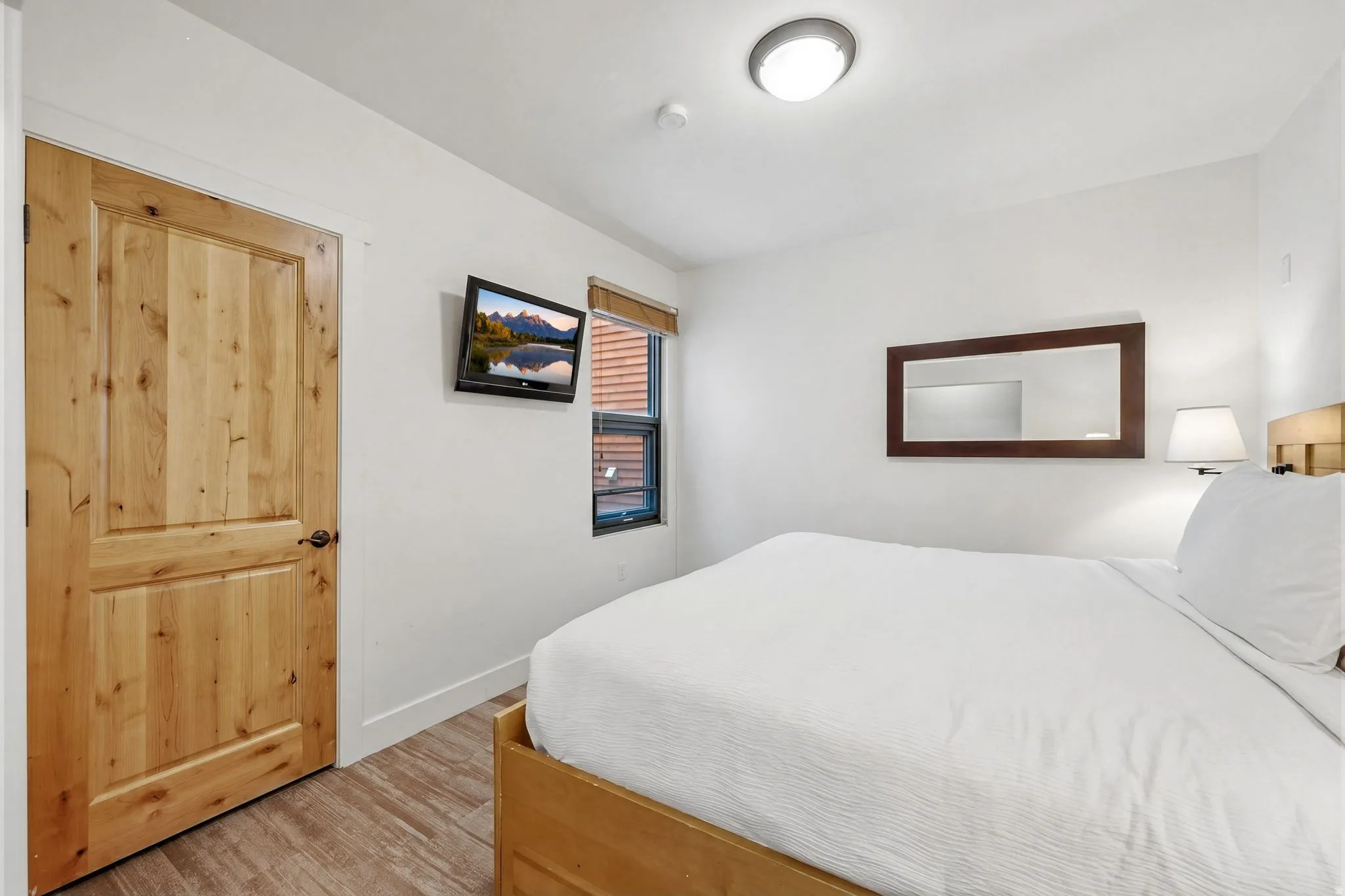 Wood-finish flooring, solid wood panel door, mounted television, wood-framed mirror, and a window with blinds