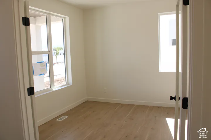 Room with light wood-finish flooring, two large windows, white walls, and a white door with black hardware
