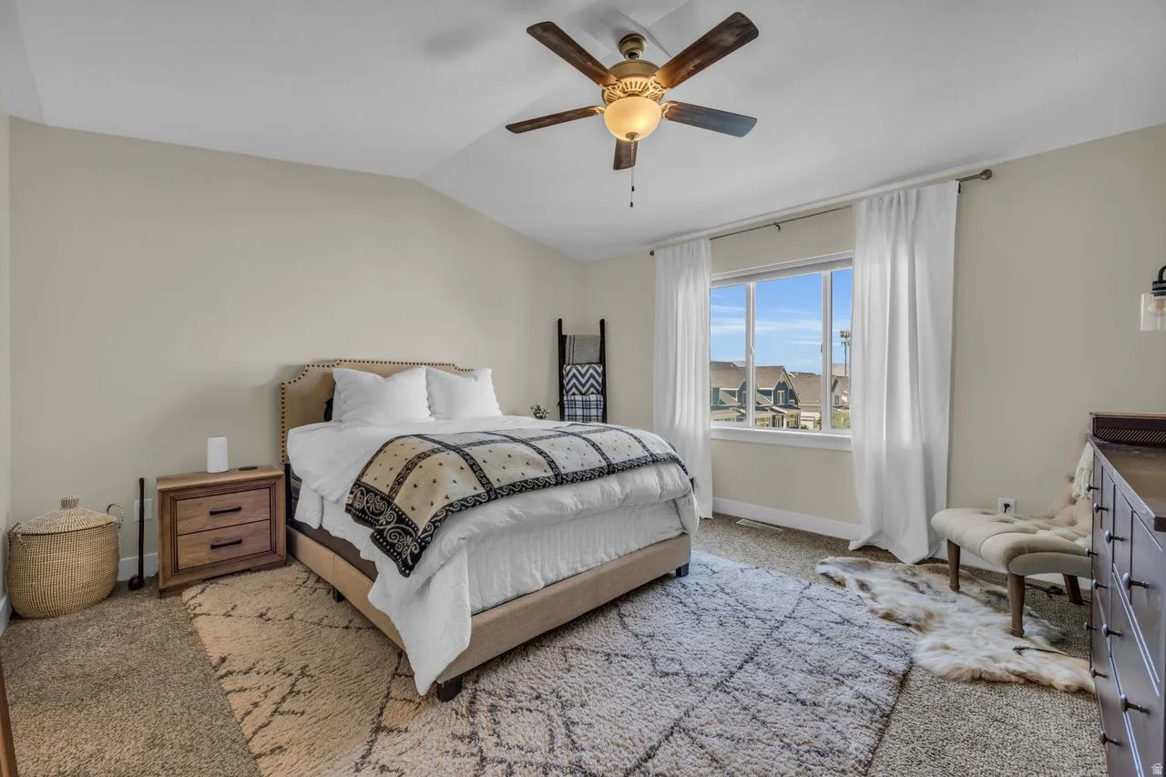 Vaulted ceiling, ceiling fan with integrated lighting, large window with white trim, wall-mounted light fixture, carpeted flooring