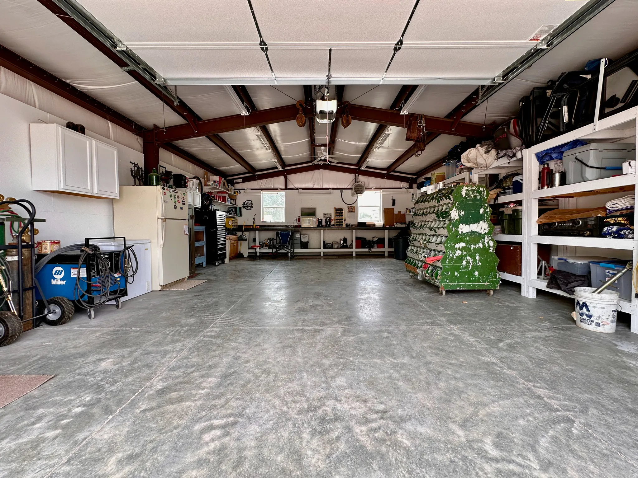 Expansive concrete floor in a workshop space, featuring exposed structural beams, white wall cabinetry, and built-in shelving units some  will stay, west wall of shelving is not included.  also the metal cover over the work bench will not stay.  It will b
