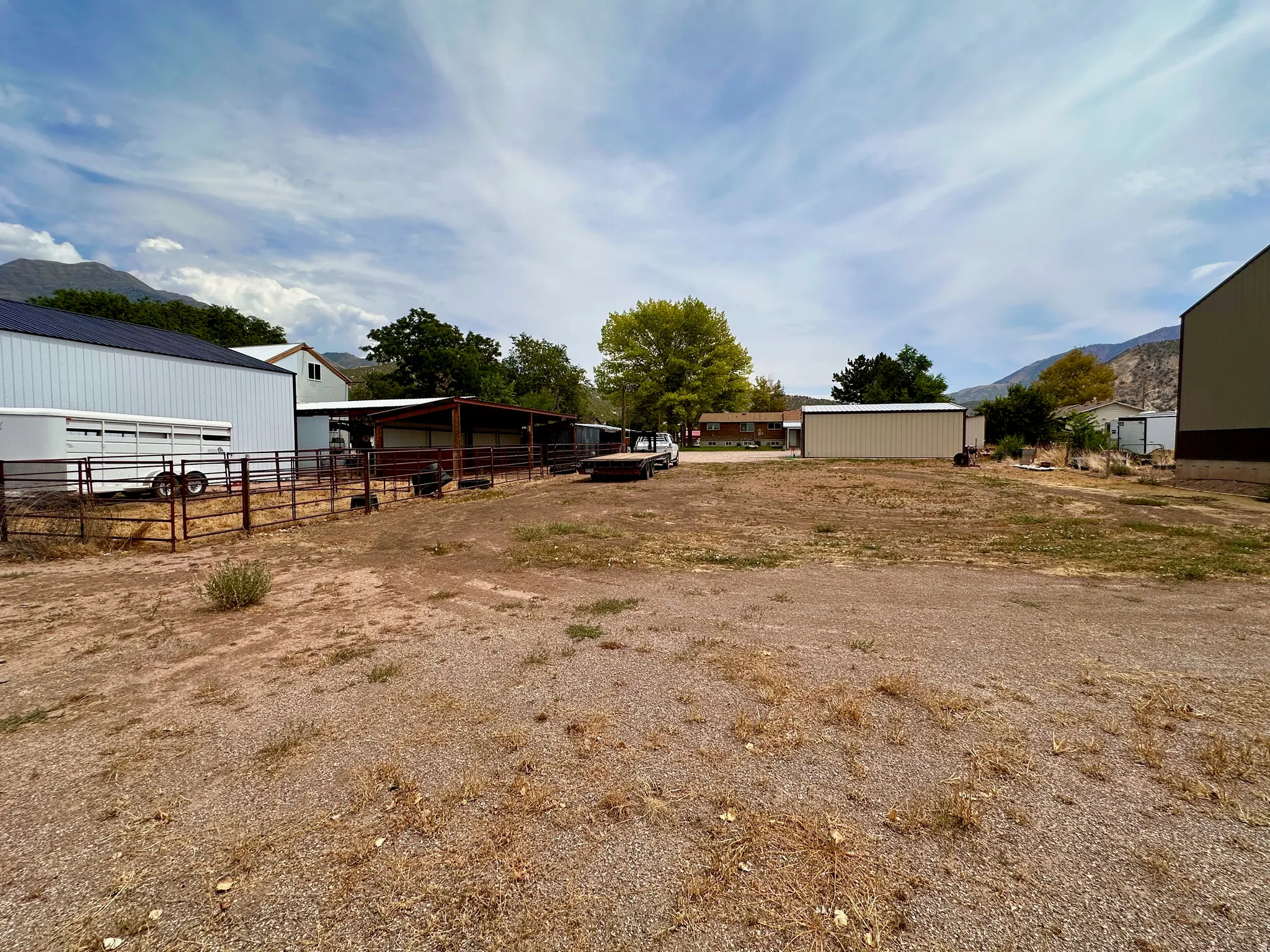 Expansive gravel and dirt lot featuring multiple metal outbuildings, a covered structure, and a fenced enclosure