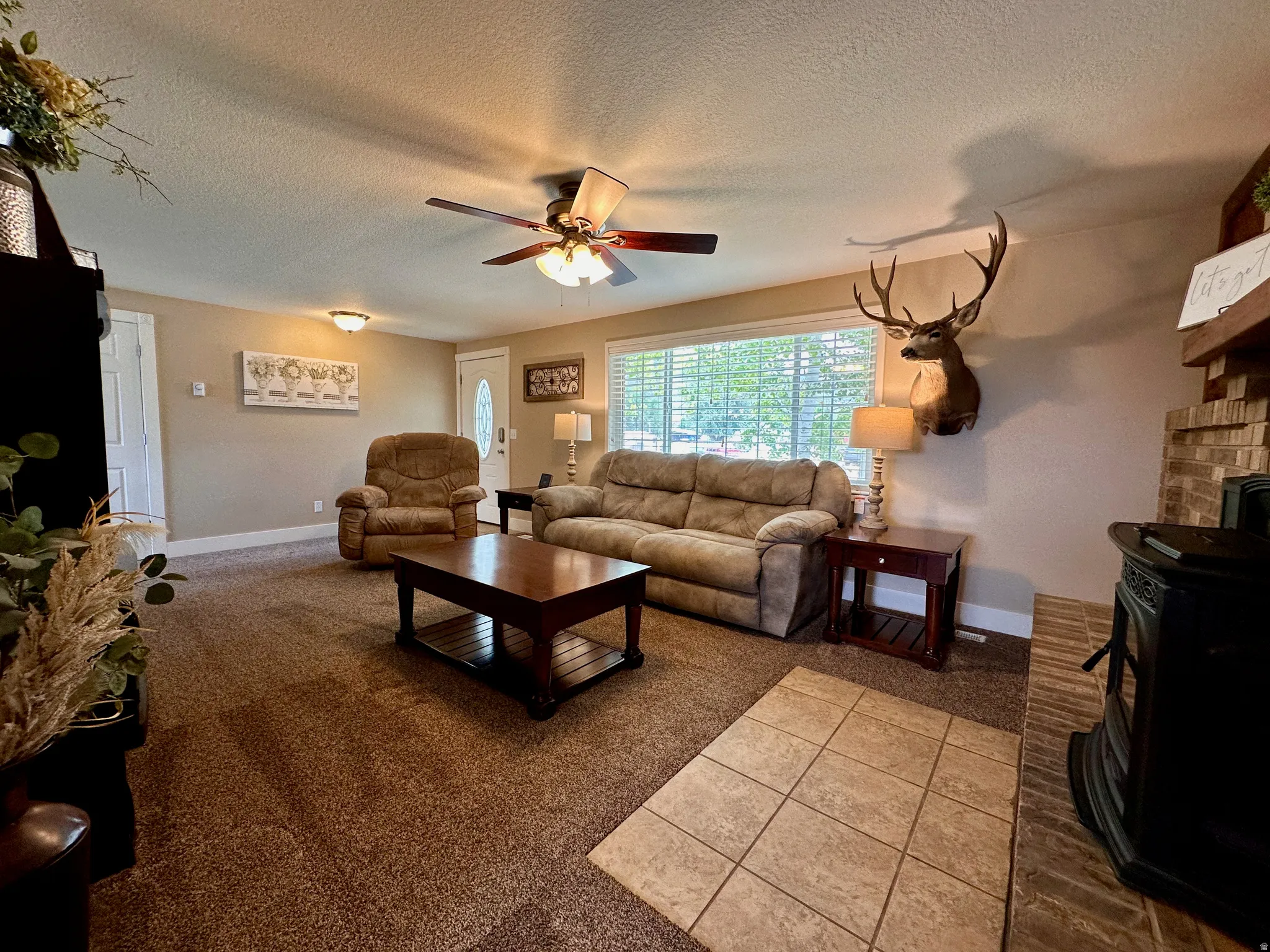 Living area featuring a wide window, ceiling fan with integrated lighting, and a brick-detailed fireplace with a wood-burning stove
