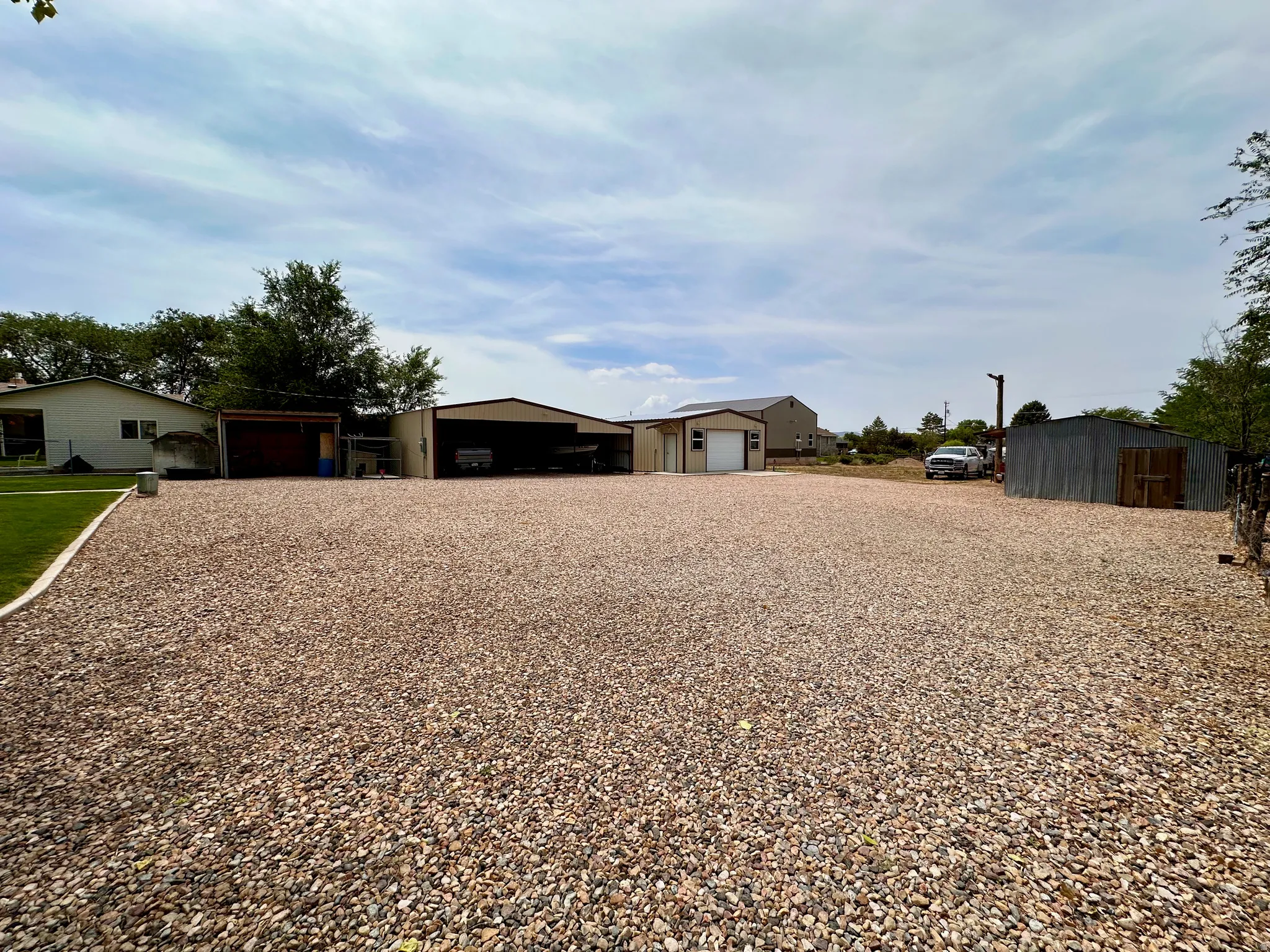 Expansive gravel lot with multiple outbuildings, including a metal-sided garage with a roll-up door, an open-sided carport, and an additional storage shed