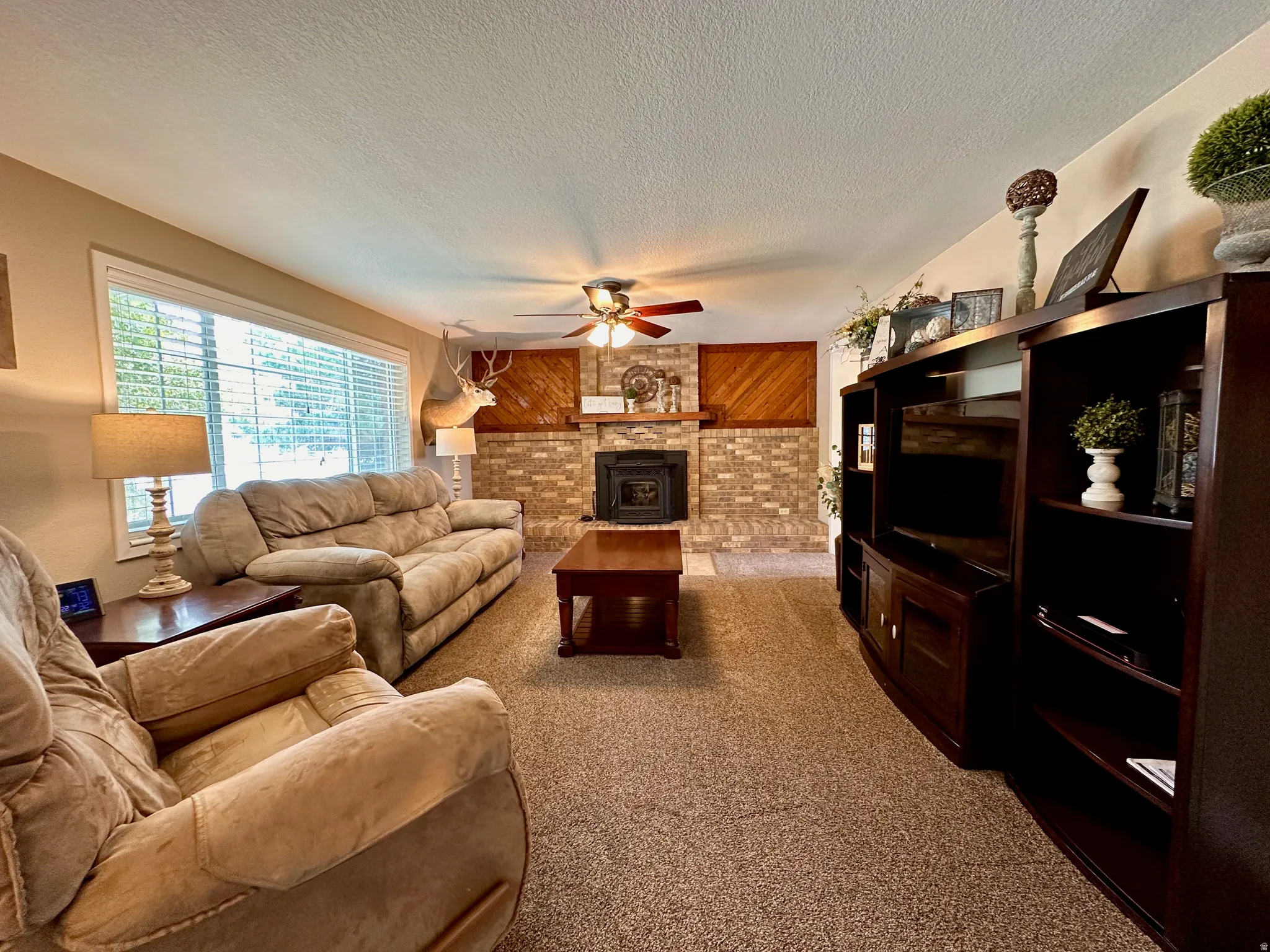 Brick fireplace with hearth, wood-paneled wall accents, ceiling fan with light fixture, and broad window with blinds