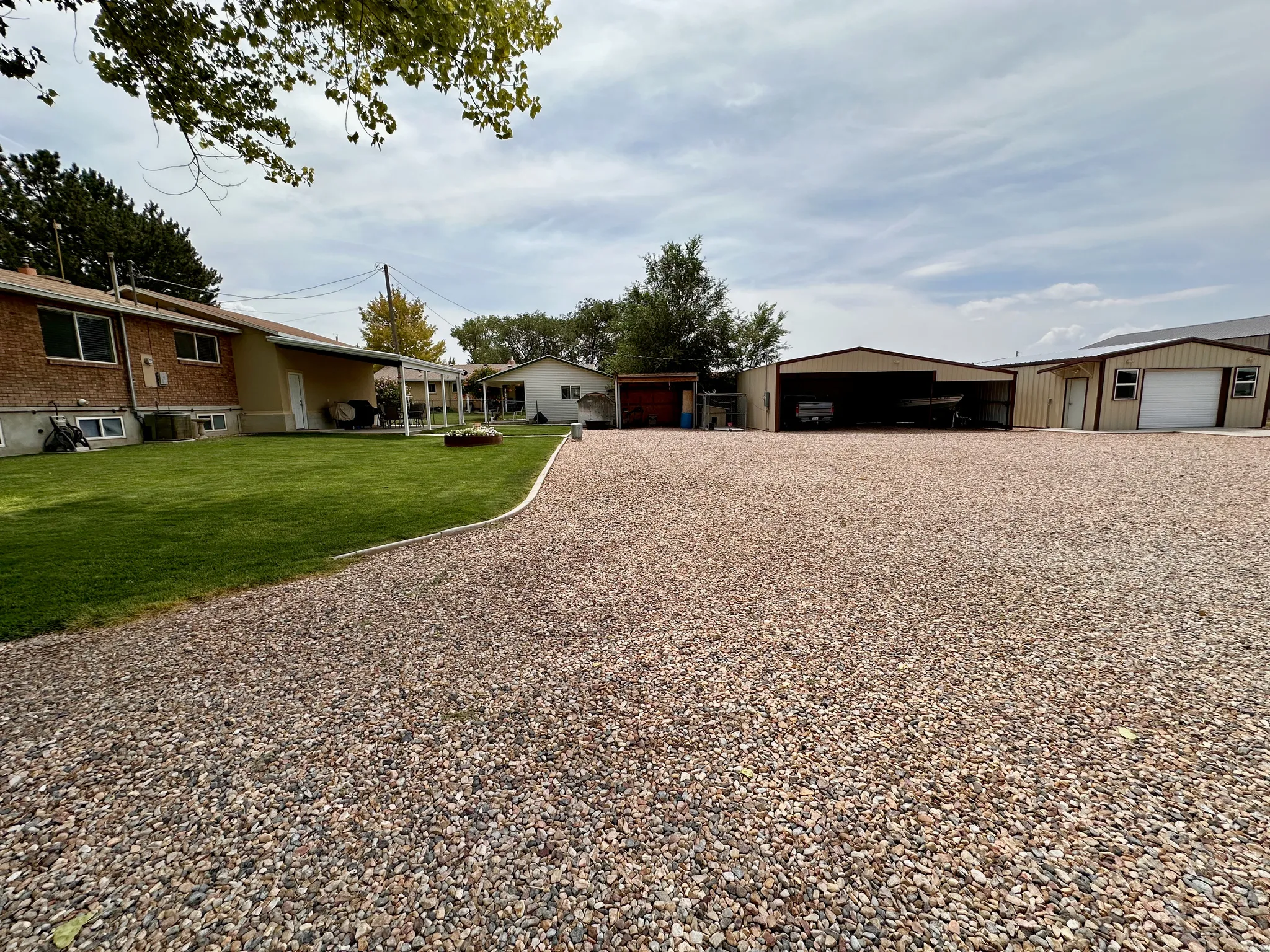 Expansive gravel area with multiple metal outbuildings, a covered patio, a manicured lawn, and a brick exterior