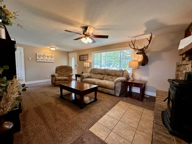 Living area featuring a wide window, ceiling fan with integrated lighting, and a brick-detailed fireplace with a wood-burning stove
