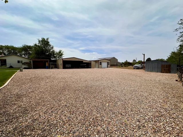 Expansive gravel lot with multiple outbuildings, including a metal-sided garage with a roll-up door, an open-sided carport, and an additional storage shed