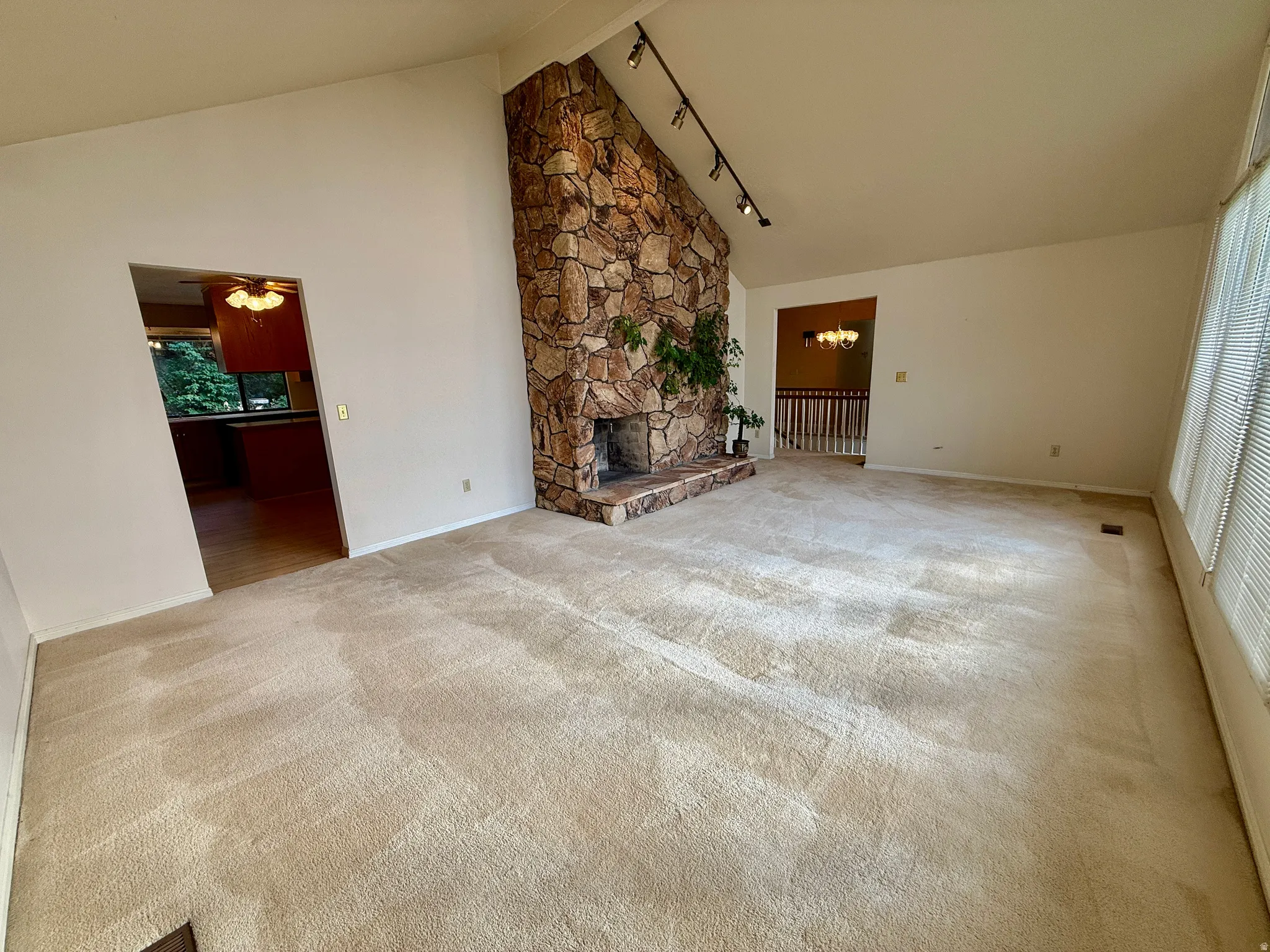 Vaulted ceiling with track lighting, stone fireplace with a raised hearth, and large windows with blinds
