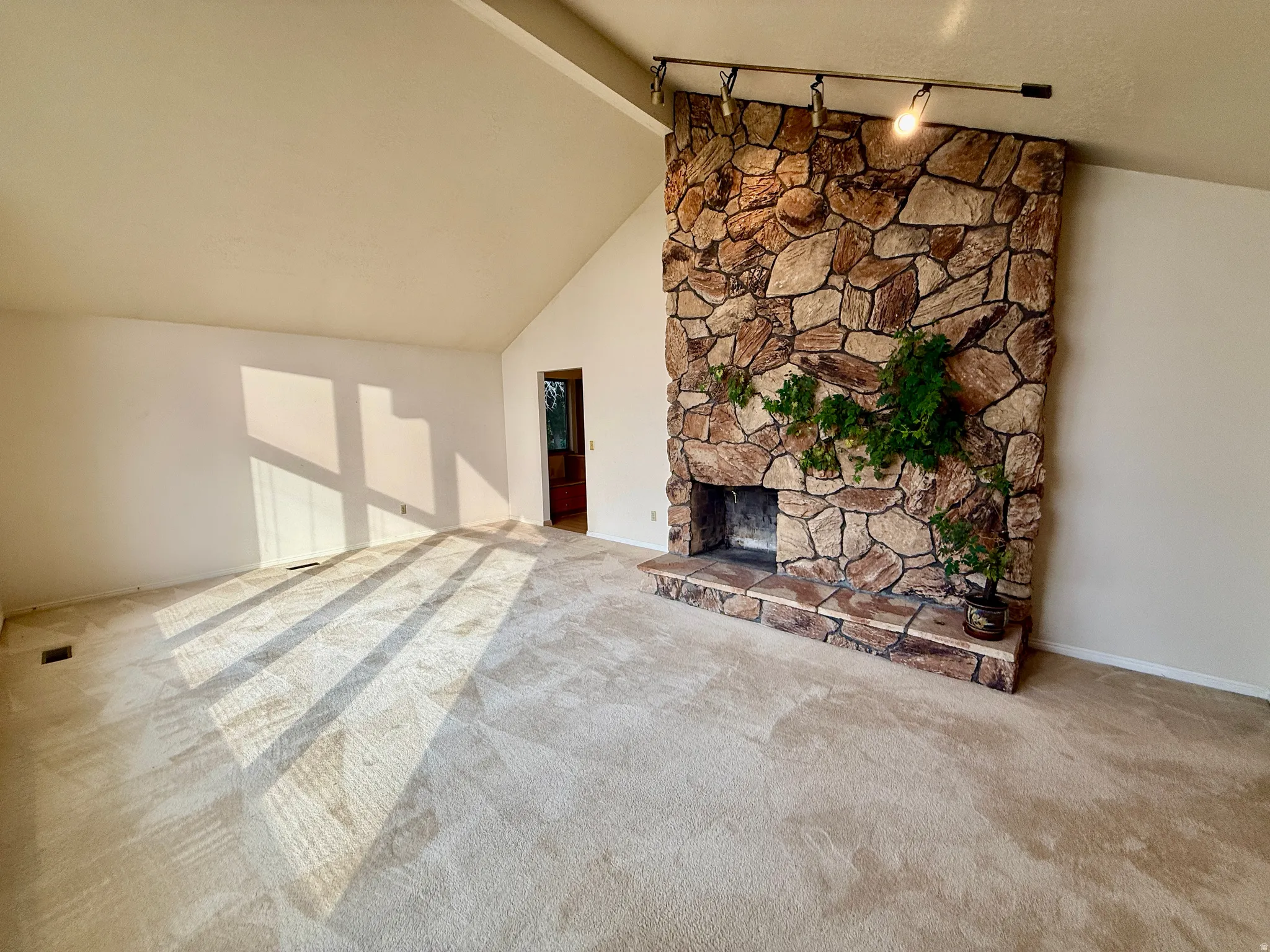 Vaulted ceiling, stone fireplace with hearth, track lighting, and neutral carpeting