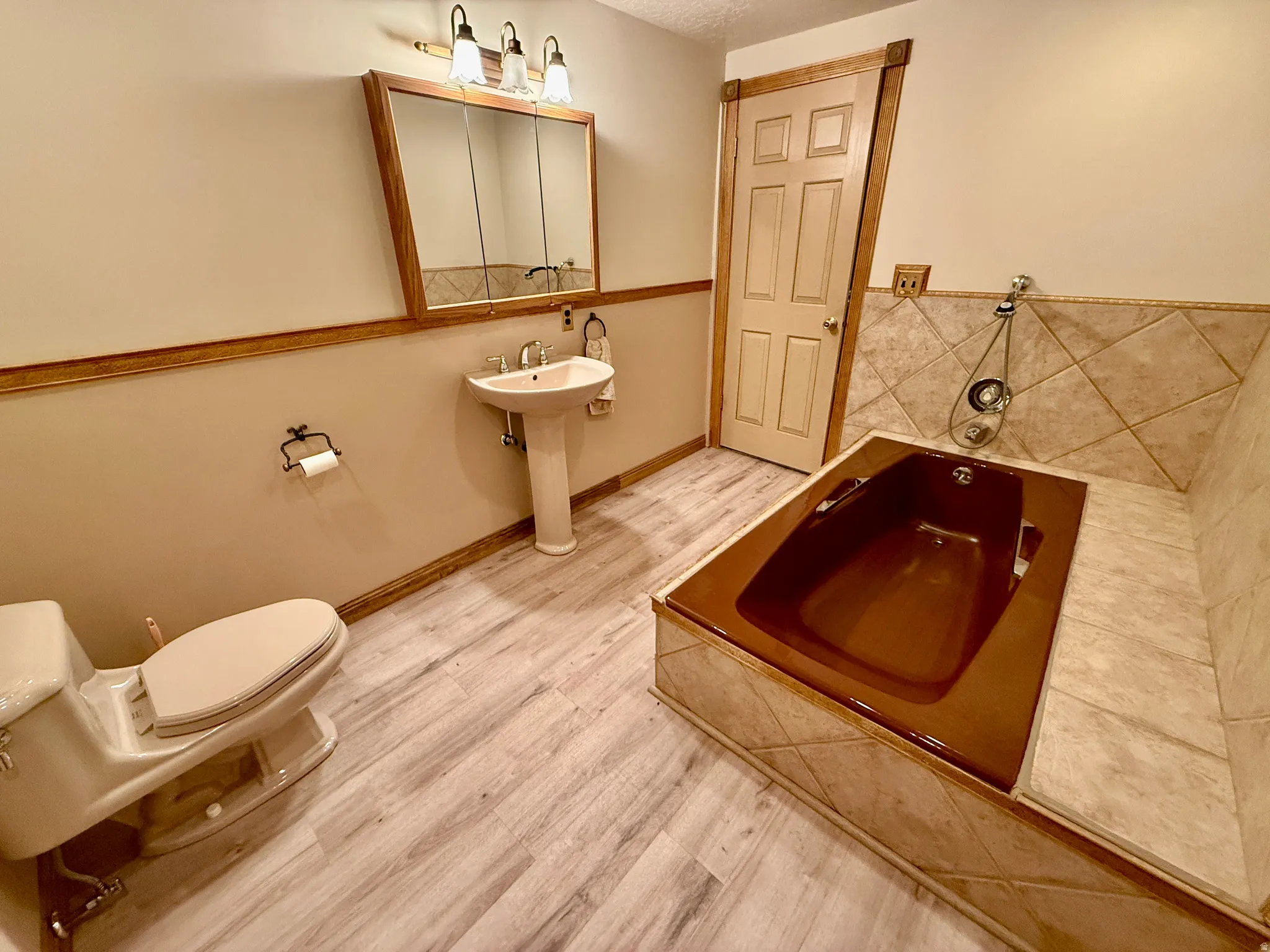 Pedestal sink with chrome fixtures, wood-finish flooring, built-in bathtub with tiled surround, wood-trimmed medicine cabinet, and three-light vanity fixture