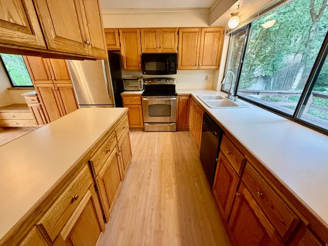Kitchen featuring wood-finish cabinetry, light-toned countertops, stainless steel appliances, a dual basin sink, and a large window overlooking natural scenery