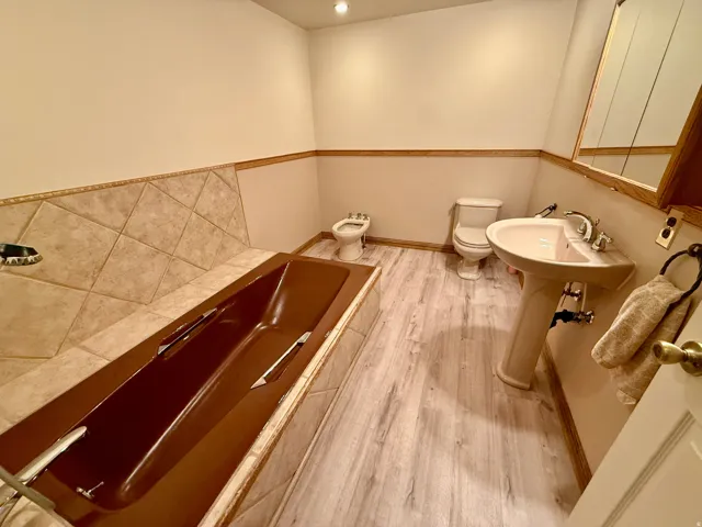 Integrated soaking tub with tiled surround, wood-finish flooring, pedestal sink, and Bidet and built-in medicine cabinet