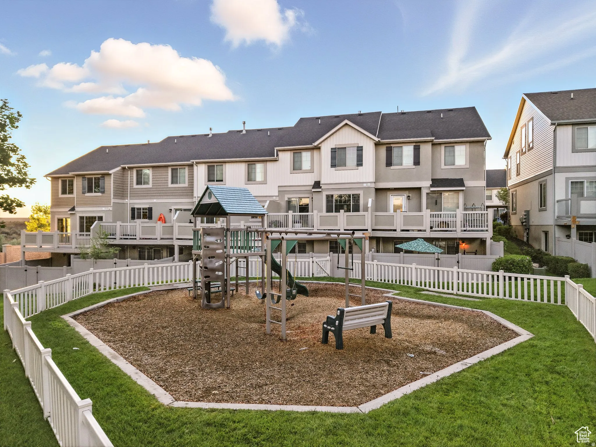 Community playground with a swing set, slide, and bench, enclosed by a white picket fence, bordered by manicured lawn