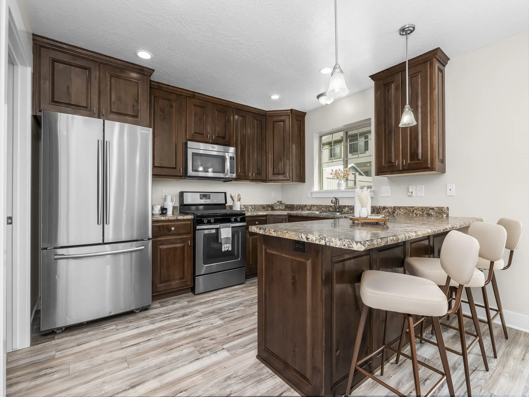 Kitchen featuring wood-finish flooring, dark wood cabinetry, stainless steel appliances, speckled countertops, and an integrated island with pendant lighting