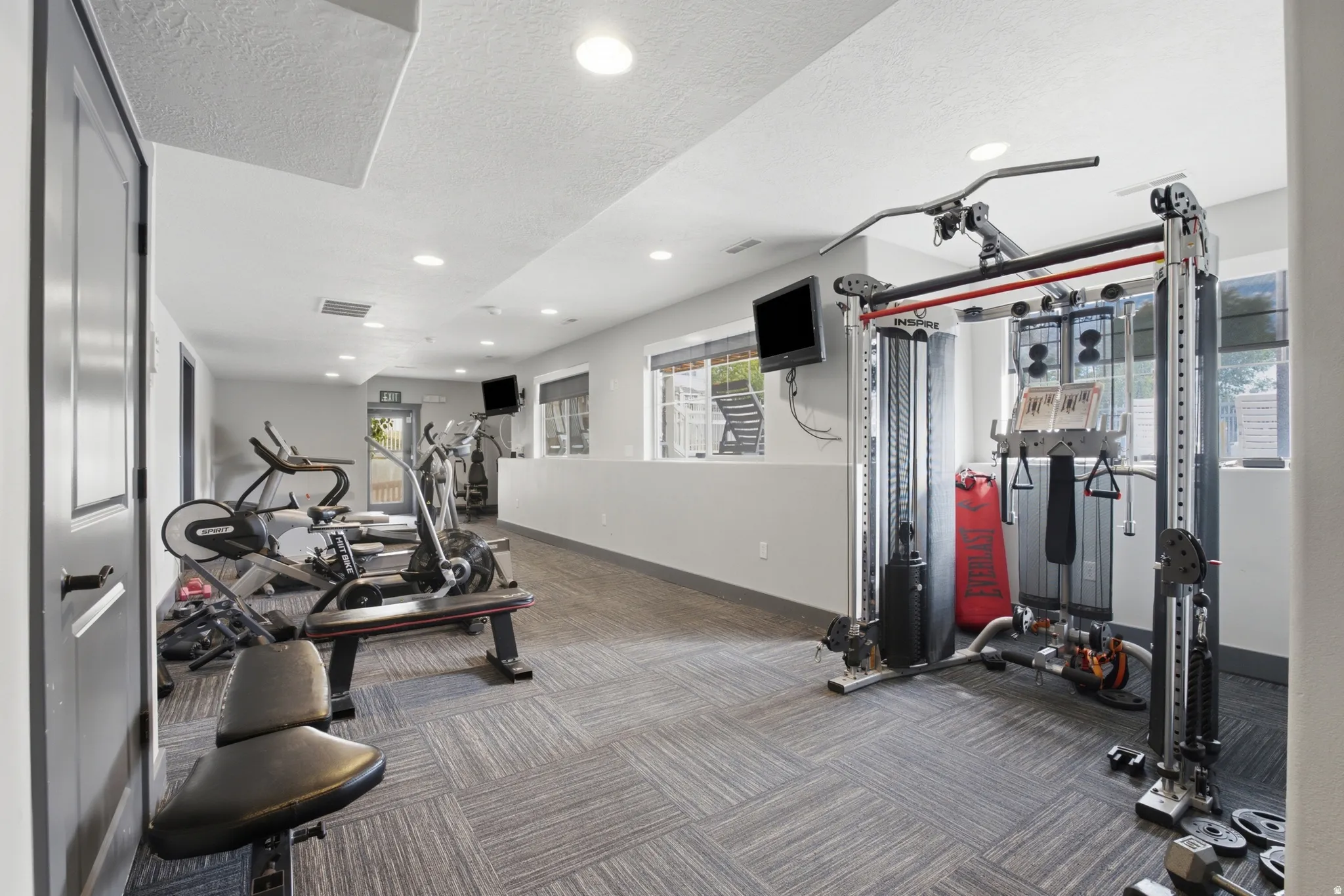 Recessed lighting illuminates the carpeted floor throughout this spacious exercise area, featuring multiple large windows and a white partition wall