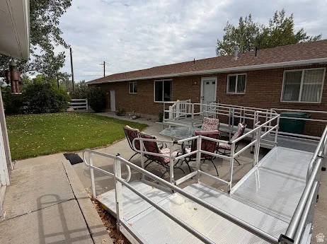 Rear exterior featuring brick siding, a brown shingle roof, concrete patio, and an accessibility ramp
