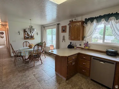 Tiled flooring, wood cabinetry, stainless steel dishwasher, window with outdoor scenery, and a chandelier-style light fixture