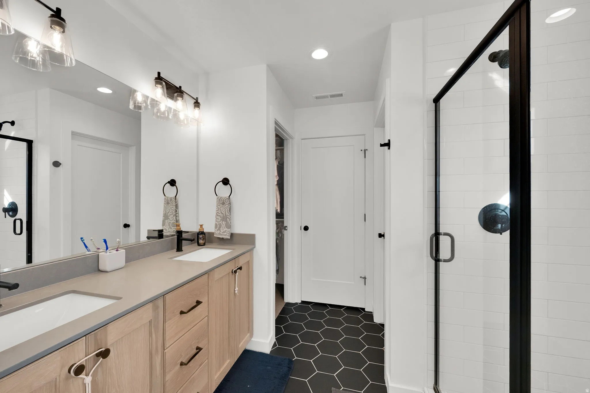 Primary bathroom featuring dual vanities with wood-finish cabinetry and gray quartz countertops, polished nickel hardware, recessed ceiling lighting, and a spacious walk-in shower with black-framed glass enclosure and hexagonal tile flooring