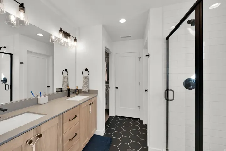 Primary bathroom featuring dual vanities with wood-finish cabinetry and gray quartz countertops, polished nickel hardware, recessed ceiling lighting, and a spacious walk-in shower with black-framed glass enclosure and hexagonal tile flooring