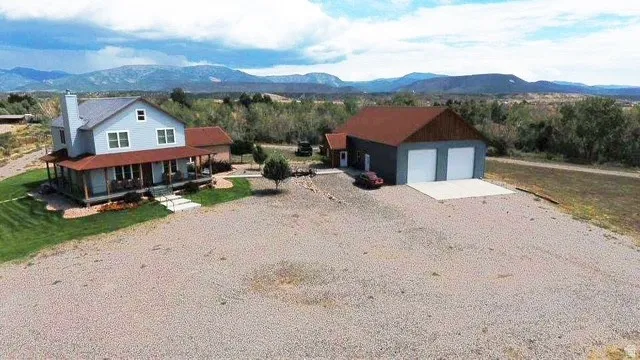 Light blue siding with white trim, covered wrap-around porch, gravel driveway, detached two-car garage with concrete pad, and distant mountain scenery