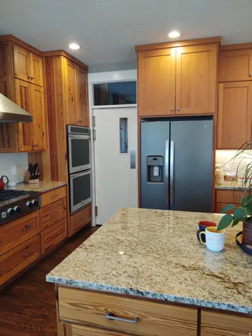 Granite island countertop with natural wood-grain cabinetry, stainless steel double wall oven, and built-in stainless steel refrigerator