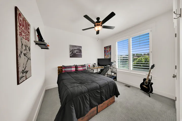 Recessed lighting, ceiling fan with integrated light fixture, dual windows with plantation shutters, wood-finish bed frame, and light gray carpet