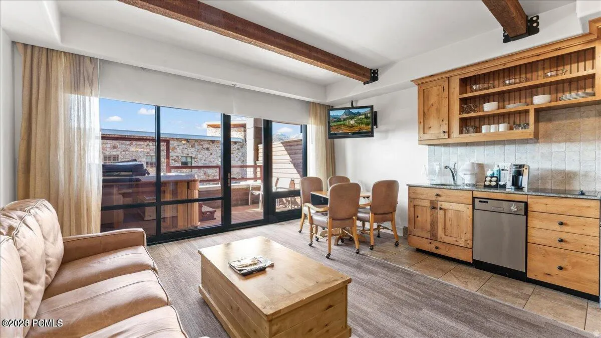 Open-concept living space with exposed wood beams, wood-finish flooring, and a full wall of glass doors leading to a private outdoor area with a built-in hot tub