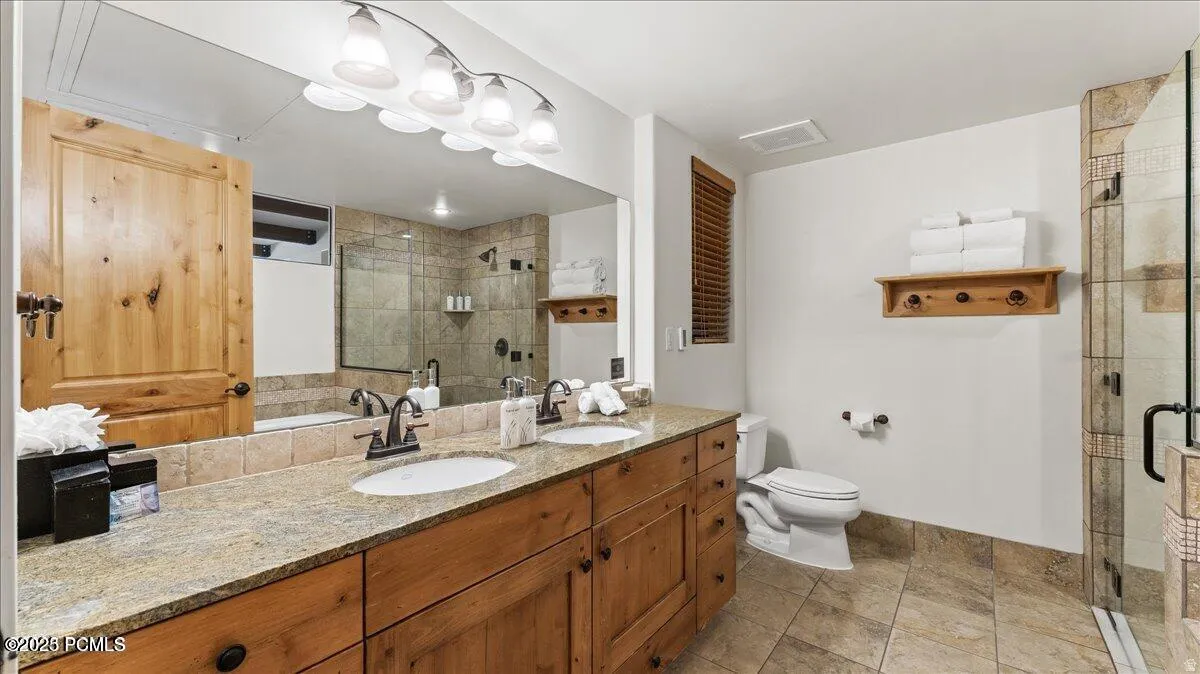 Dual vanity with stone countertop, wood-finish cabinetry, tile flooring, and a glass-enclosed shower